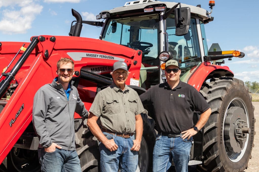 Three men posing in front of a large red Massey Ferguson tractor on a sunny day with blue sky and white clouds behind them.