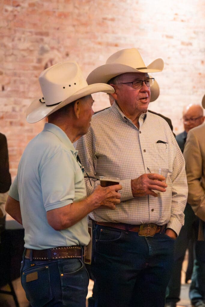 Two older men wearing cowboy hats and glasses converse at a social event, each holding a drink.