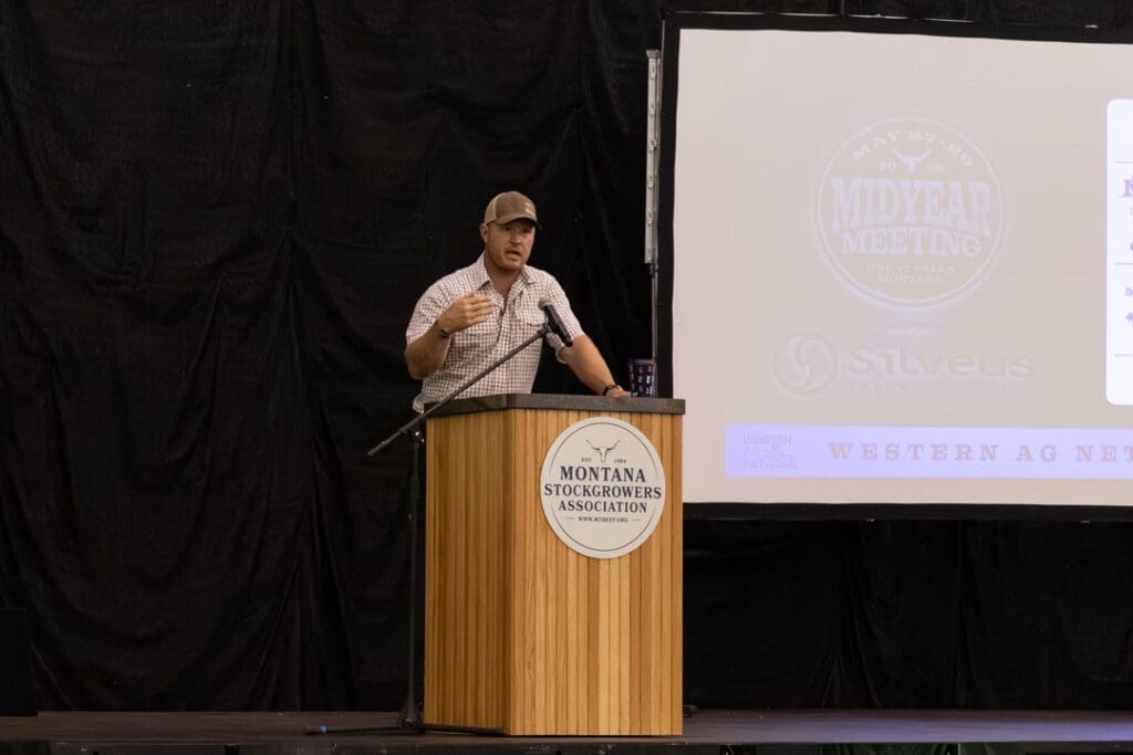 Man at a wooden podium giving a talk at a Montana Stockgrowers Association event, with a projection screen to the right.