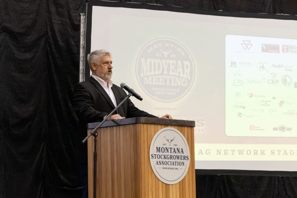 Man in a dark suit speaks at a wooden podium with a Montana Stockgrowers Association emblem; a projection screen shows 'Midyear Meeting' behind him.
