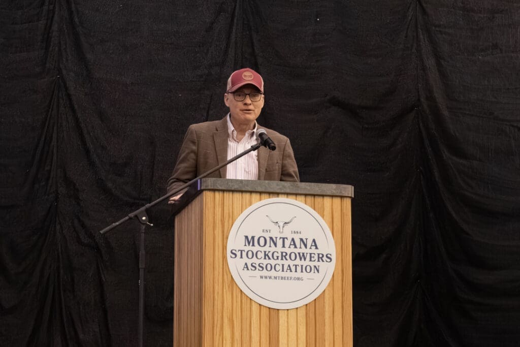 Man wearing glasses and a maroon cap speaks at a wooden podium with the Montana Stockgrowers Association logo.