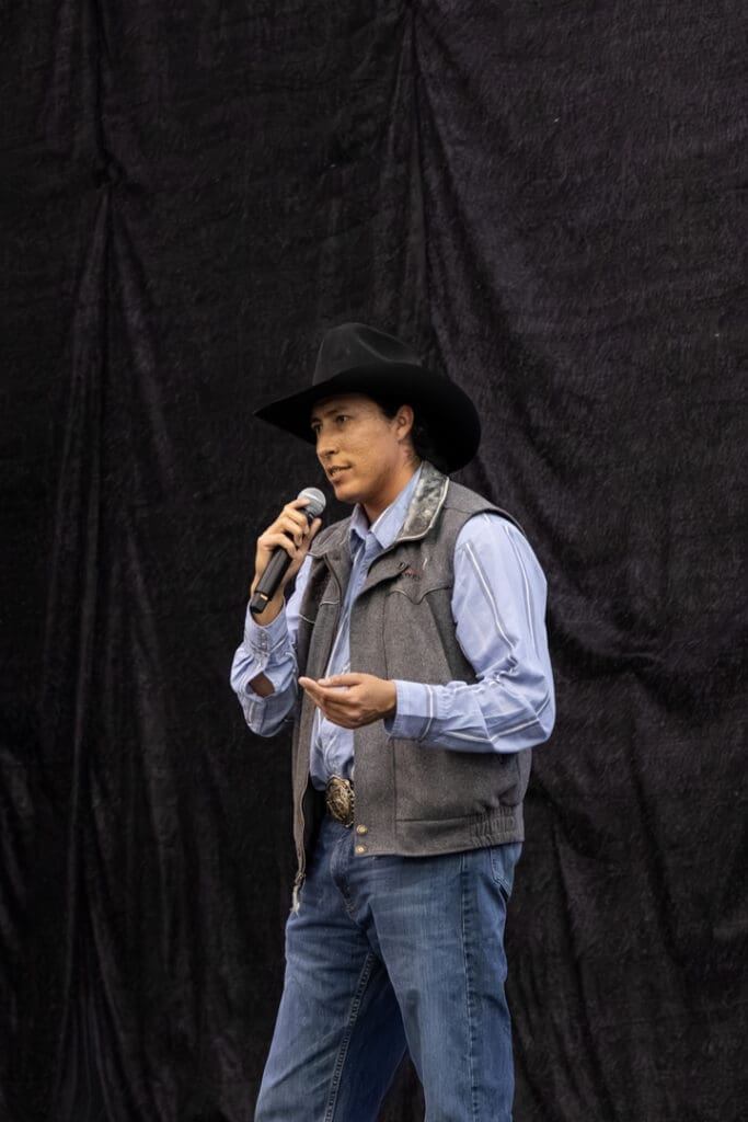 Man wearing a cowboy hat and gray vest speaks into a microphone on a dark stage backdrop.
