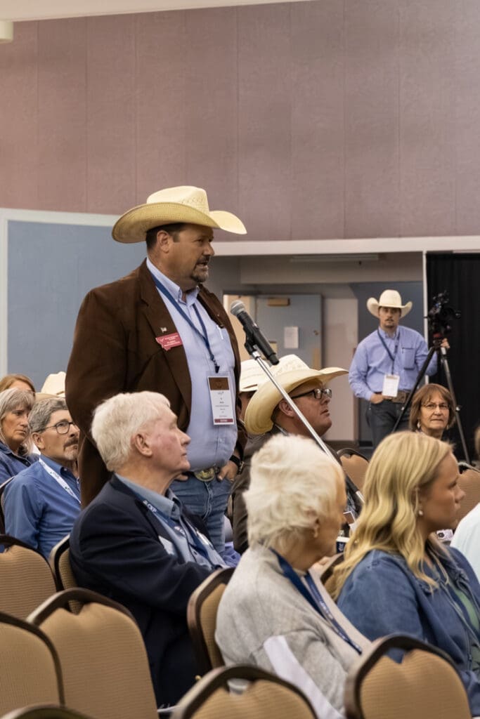 Man wearing a cowboy hat speaks into a microphone at a conference, addressing a seated audience.