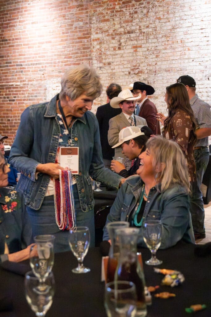 Two women in denim jackets laugh as one hands red, white, and blue bead necklaces to a seated guest at a casual party, brick wall behind them.