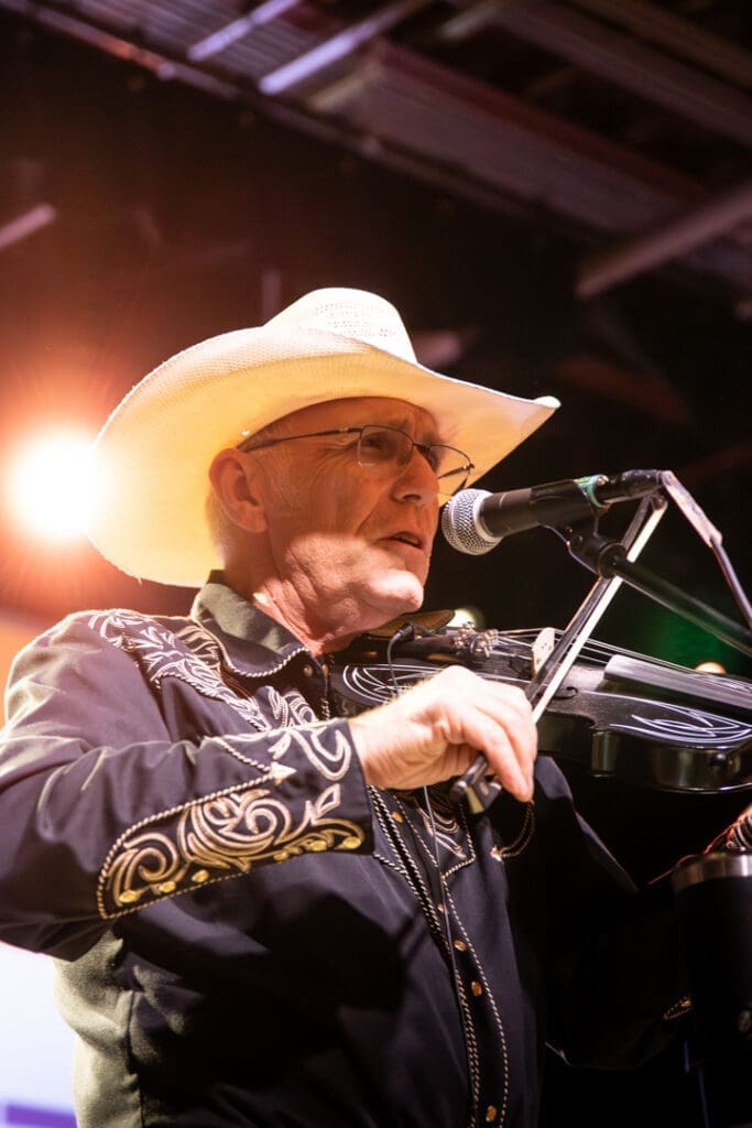 Older man in a white cowboy hat playing a violin on stage, singing into a microphone during a live performance.