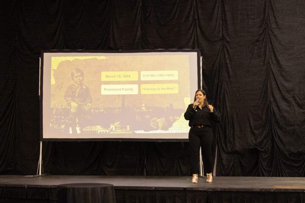 Female presenter on a stage speaking into a microphone, next to a projected slide with a vintage cityscape and yellow labeled boxes about 1864 and the Civil War.