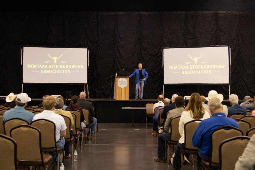 Speaker on stage at a podium addressing an audience at a Montana Stockgrowers Association event.